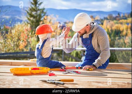 Padre con bambino figlio costruzione legno cornice casa. Costruttore maschile che dà alto cinque a capretto in cantiere, indossare casco e tute blu in giornata di sole. Carpenteria e concetto di famiglia. Foto Stock