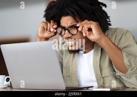 Concentrato giovane ragazzo nero riccio in bicchieri squints e guarda il portatile a tavola con tazza in interno caffè Foto Stock