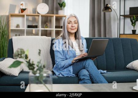 Piacevole donna dai capelli grigi con lunghi capelli seduti sul divano con computer portatile in ginocchio. Felice pensionato femmina godendo di lavoro remoto da casa. Concetto di pensionamento e tempo libero. Foto Stock