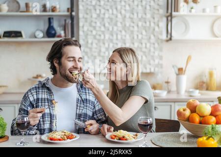 Allegra giovane donna europea nutre il marito di spaghetti con verdure o pasta in una cucina minimalista Foto Stock