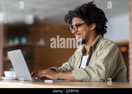 Felice giovane africano americano curly maschio in occhiali lavora al portatile al tavolo con tazza in caffè interno, profilo Foto Stock