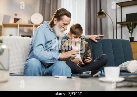 Buon nonno senior e simpatico nipote utilizzando le app per smartphone a casa, nonno vecchio che abbraccia nipote tenendo in mano lo schermo del cellulare giocare gioco mobile relax sedersi sul divano Foto Stock