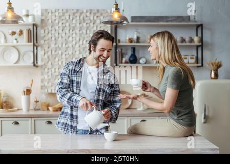 Felice giovane moglie europea e marito con tobble drink tè caldo e parlare nel tempo libero in cucina moderna Foto Stock