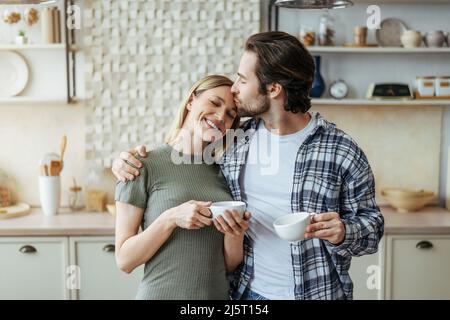 Soddisfatto giovane uomo caucasico con baci stoppia moglie sulla fronte bere caffè in cucina minimalista interno Foto Stock