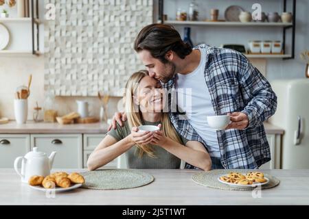 Soddisfatto giovane europeo uomo con stoppia baci donna, bere bevanda calda nel tempo libero in cucina moderna interno Foto Stock
