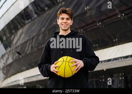 Ritratto di giovane uomo sorridente in felpa nera con cappuccio e palla da basket. Foto Stock