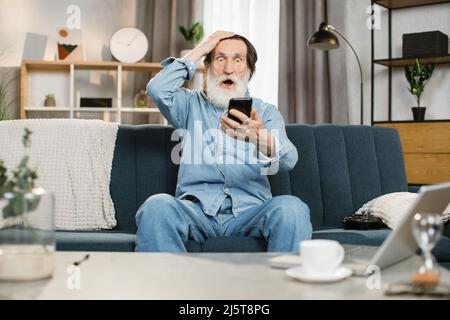 Pensionato caucasico con barba grigia con viso emozionale felice e pugno sollevato dopo aver ricevuto buone notizie sullo smartphone a casa leggere notizie mentre si rilassa sul comodo divano blu in soggiorno Foto Stock