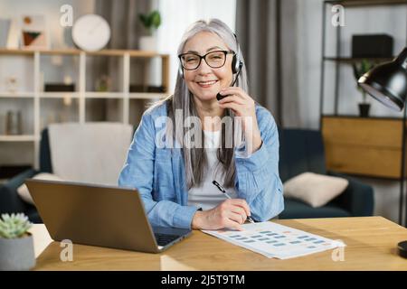 Donna anziana positiva che utilizza cuffie e laptop per le conversazioni video in ufficio. Donna anziana freelancer seduta a tavola e prendere appunti durante la video chat. Foto Stock