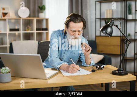 Vista frontale dell'uomo maturo duro con l'investitore professionale della barba grigia, socio che fuma sigaro seduto al tavolo in ufficio moderno mentre controlla i documenti del contratto d'affari. Foto Stock