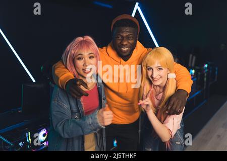 Tre allegri amici interrazziali - uomo afroamericano, donne asiatiche e caucasiche - guardando la macchina fotografica, e sorridendo, mentre si trova in piedi in un bar di gioco. Foto di alta qualità Foto Stock