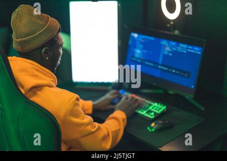 Programmatore freelance maschile vestito casualmente afroamericano seduto in uno studio, cliccando sulla tastiera, guardando lo schermo del computer. Foto di alta qualità Foto Stock