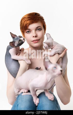 Giovane donna rossa che guarda la macchina fotografica, tenendo due Sphynx Cat nelle mani e un gattino seduto sulle gambe. Donna carina con capelli corti Foto Stock