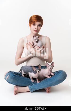 Giovane donna rossa che tiene Sphynx Cat nelle mani e due gattini seduti sulle gambe. Donna Hipster con capelli corti in T-shirt e jeans guardare macchina fotografica Foto Stock