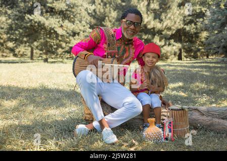 Bell'uomo africano in costume nazionale suona un tamburo etnico, djembe insieme a figlio. Foto Stock