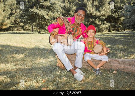 Bell'uomo africano in costume nazionale suona un tamburo etnico, djembe insieme a figlio. Foto Stock