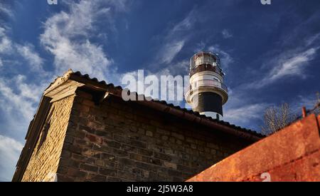 faro con parte di case . Su cielo blu con nuvole. Torre a luci rosse. Foto Stock