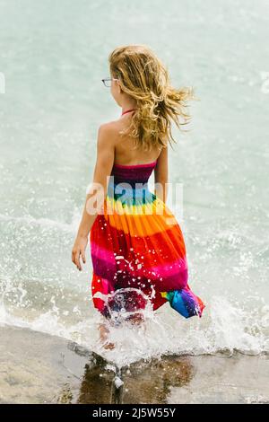 La ragazza bionda si alza in piedi nell'oceano mentre il vento soffia i capelli e le onde colpiscono le gambe Foto Stock