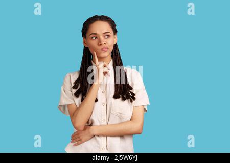 Ritratto di donna pensierosa con dreadlocks neri pensando al futuro, tenendo il mento, avendo espressione facciale seria, indossando la camicia bianca. Studio interno girato isolato su sfondo blu. Foto Stock