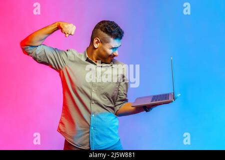 Uomo arrabbiato in camicia che punisce lo schermo del laptop, guardando con furiosa pazza espressione, boxe minacciando di colpire mentre si fa videochiamata. Studio interno girato isolato su colorato sfondo di luce al neon. Foto Stock