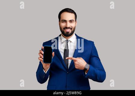 Uomo bearded che punta il dito verso lo smartphone con schermo vuoto, guardando la fotocamera con sorriso, spazio libero per la pubblicità, indossando abito stile ufficiale. Studio interno girato isolato su sfondo grigio. Foto Stock