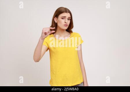 Per favore, un po' di più. Ritratto di ragazza adolescente carina in T-shirt gialla che mostra piccolo formato o piccolo gesto della punta con le dita, guardando con implorazione grimace. Studio interno girato isolato su sfondo grigio. Foto Stock