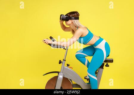 Ritratto laterale della donna grinning che guida il simulatore di bicicletta con la realtà virtuale cuffia, facendo cardio addestramento, indossando il blu sportswear. Studio interno girato isolato su sfondo giallo. Foto Stock