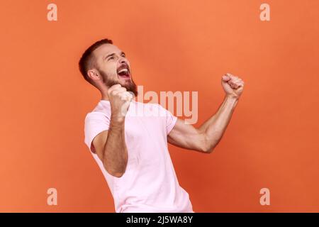 Ritratto di un bell'uomo strariciato che esprime il gesto vincente con i pugni rialzati e urla, celebrando la vittoria, indossando una T-shirt rosa. Studio interno girato isolato su sfondo arancione. Foto Stock
