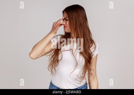 Ritratto di donna dispiaciuta confuso con capelli scuri naso pizzicando per evitare cattivo odore, esprimendo repulsione e disgusto, indossando T-shirt bianca. Studio interno girato isolato su sfondo grigio. Foto Stock