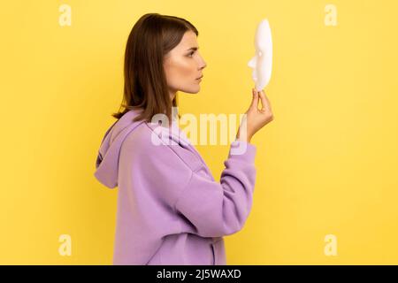 Profilo di giovane adulta attraente donna che tiene la maschera bianca nelle mani, personalità nascosta, esprimendo calma emozioni severe, indossando felpa con cappuccio viola. Studio interno girato isolato su sfondo giallo. Foto Stock