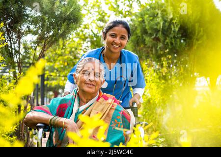 Infermiera sorridente felice che prende la donna anziana a piedi mentre sulla sedia a rotelle al giardino dell'ospedale guardando la macchina fotografica - concetto di custode, invalidità e. Foto Stock