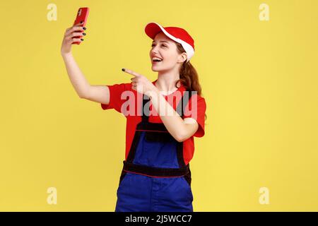 Ritratto di felice sorridente lavoratore donna che trasmette in diretta streaming e puntando il dito verso la fotocamera del telefono cellulare, indossando tute e berretto rosso. Studio interno girato isolato su sfondo giallo. Foto Stock