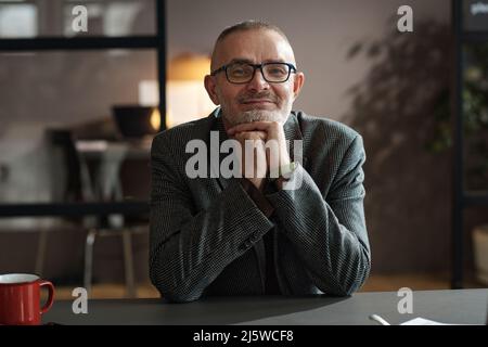 Ritratto di uomo d'affari maturo in occhiali guardando soddisfatto alla macchina fotografica mentre si siede alla scrivania in ufficio Foto Stock