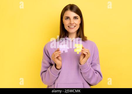 Donna sorridente che tiene e mostra due puzzle pezzi, matching e collegare puzzle, risolvere problemi con buon risultato, indossare felpa con cappuccio viola. Studio interno girato isolato su sfondo giallo. Foto Stock