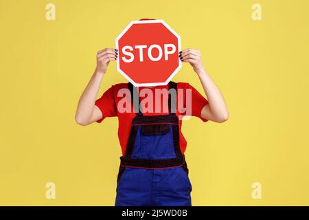 Ritratto di donna operaia in piedi nascondendo il suo volto dietro un segno rosso di stop, divieti e restrizioni, indossare tute e cappuccio rosso. Studio interno girato isolato su sfondo giallo. Foto Stock