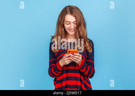 Donna felice indossando a righe stile casual maglione, con piacevole sorriso utilizzando il telefono cellulare, rispondere ai messaggi nei social network, scrivere post. Studio interno girato isolato su sfondo blu. Foto Stock