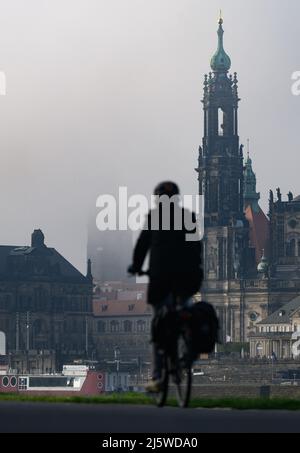 Dresda, Germania. 26th Apr 2022. La torre del municipio (l) è ancora avvolta nella nebbia al mattino, mentre un ciclista corre lungo la pista ciclabile dell'Elba di fronte alla città vecchia con la Hofkirche. Credit: Robert Michael/dpa/Alamy Live News Foto Stock