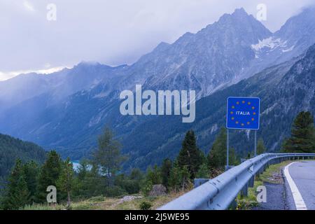 Segnale stradale al confine italiano con molte stelle gialle dell'Unione europea e paesaggio di vette della montagna Hochgall coperte da nuvole, valle e Anth Foto Stock