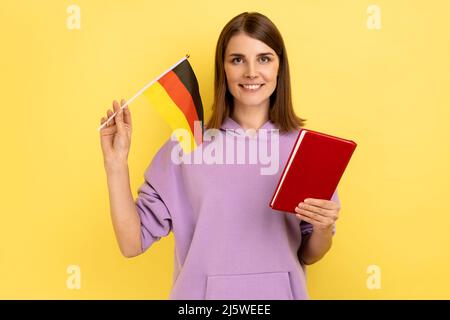 Ritratto di bella sorridente donna pelata soddisfatto tenendo libro e bandiera tedesca, istruzione all'estero in Germania, indossando felpa con cappuccio viola. Studio interno girato isolato su sfondo giallo. Foto Stock