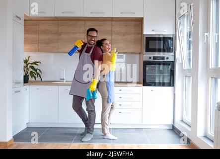 Giovane coppia in cucina con forniture di pulizia che indossano guanti e che fanno le loro faccende insieme. Foto Stock