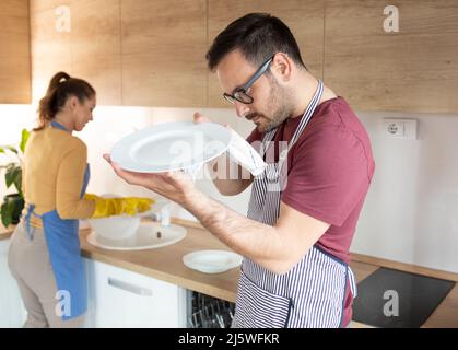 L'uomo con un grembiule in cucina tiene in mano un piatto e lo lucida con un panno mentre una donna sullo sfondo lava i piatti Foto Stock
