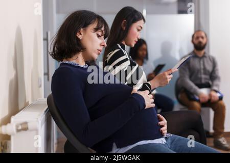 Reclutatore di lavoro incinta che tiene la mano sul ventre in attesa nell'area della lobby dell'ufficio affari di startup. Gruppo multietnico di candidati che leggono CV riprendere prima della riunione di colloquio. Concetto di reclutamento Foto Stock