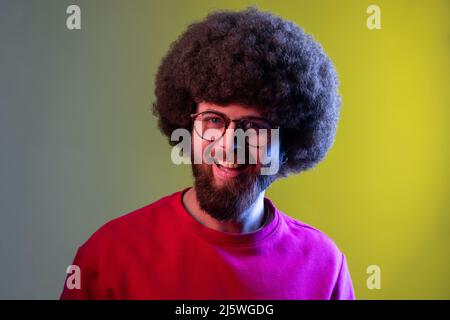 Ritratto di hipster uomo con acconciatura Afro guardando la macchina fotografica con sorriso felice, ha buon umore, indossando felpa rossa. Studio interno girato isolato su colorato sfondo di luce al neon. Foto Stock
