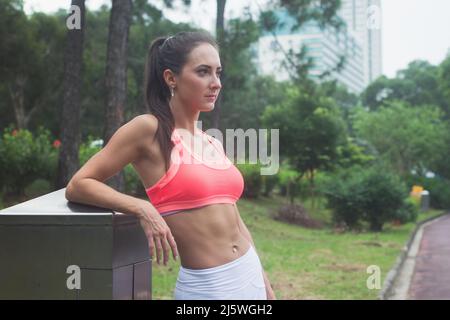 Sottile donna con bruna fitness con sei ABS che indossa un reggiseno sportivo rosa in piedi nel parco della città rilassante dopo l'allenamento guardando lontano dalla fotocamera. Foto Stock