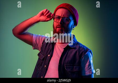 Un uomo di hipster serio che guarda la macchina fotografica con uno sguardo attento, mantenendo le braccia sulla montatura degli occhiali, indossando cappello da berretto e gilet in denim. Studio interno girato isolato su colorato sfondo di luce al neon. Foto Stock