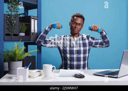 Giovane adulto attivo e sano che fa l'allenamento con manubri fitness al fine di mantenere il corpo forte. Uomo d'affari atletico azienda che solleva pesi mentre si siede alla scrivania in ufficio. Foto Stock