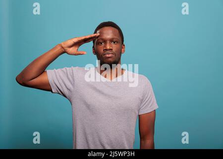 Giovane uomo che fa saluto militare con mano sulla fronte, pubblicità onore e rispetto fidato di fronte alla macchina fotografica. Orgoglioso adulto che mostra il gesto di ciao del soldato dell'esercito in studio. Saluto del veterano Foto Stock