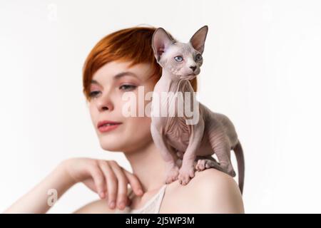 Sphynx gattino senza capelli seduto sulla spalla di una giovane donna rossa con capelli corti. Colpo di testa su sfondo bianco. Fuoco selettivo sul gatto Foto Stock