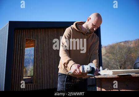 Calvo maschio operaio taglio metallo, utilizzando sega circolare mentre costruire casa telaio in legno in giorno di sole con cielo blu sfondo. Concetto di costruzione. Foto Stock