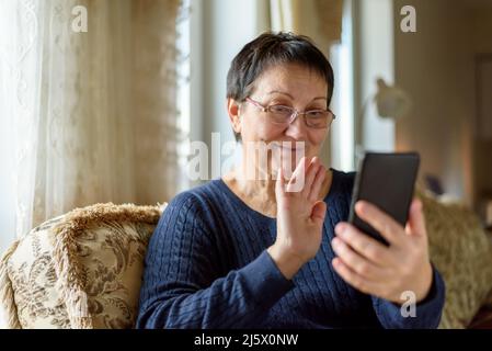 Donna anziana negli occhiali che effettua videochiamate sullo smartphone a casa Foto Stock