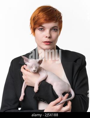 Giovane donna Bossy vestita in giacca nera che tiene le mani dormendo Sphynx kitten blu mink e bianco colore. Bella donna rossa con capelli corti Foto Stock